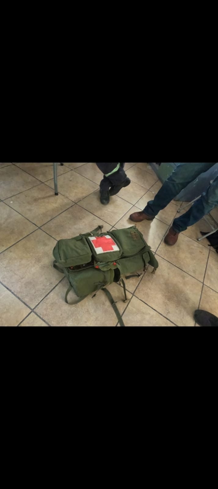 Mochila de primeros auxilios verde con cruz roja en el suelo, rodeada de personal