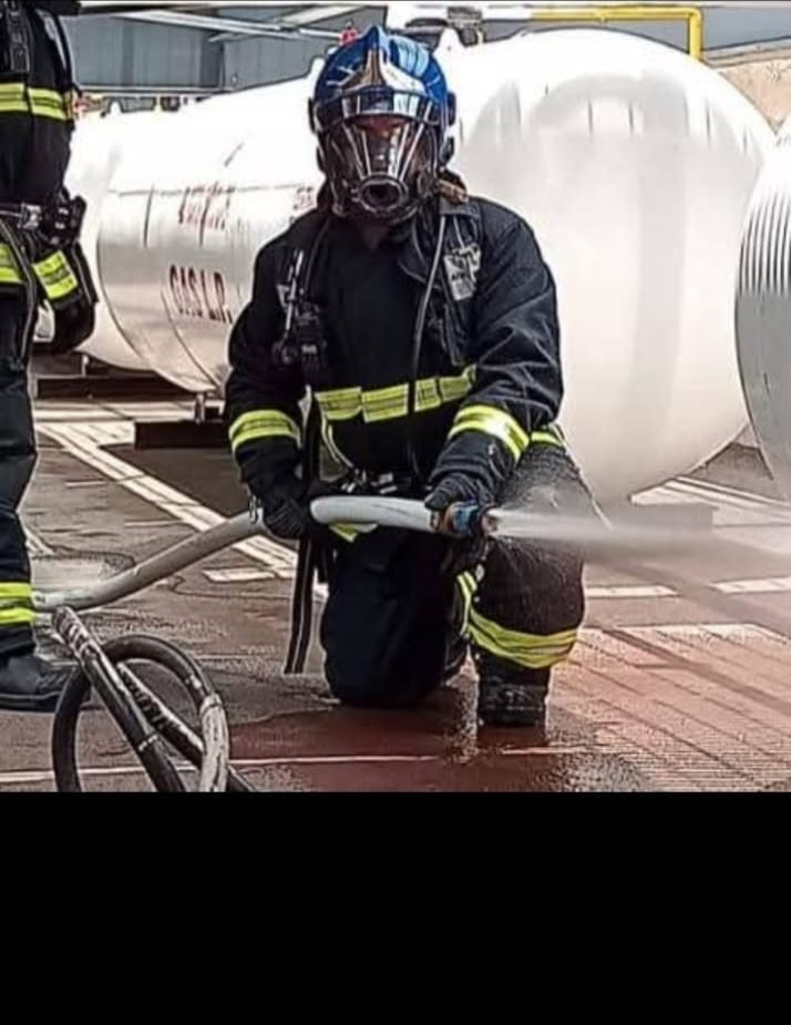 Bombero con equipo SCBA completo operando una manguera junto a un tanque industrial