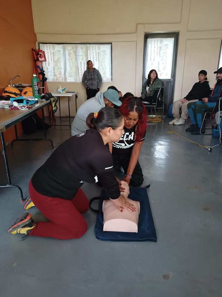 Grupo practicando RCP en maniquí adulto en un salón con equipo de rescate en altura visible al fondo