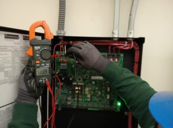 Técnico con multímetro de gancho midiendo un panel de control con tarjeta electrónica